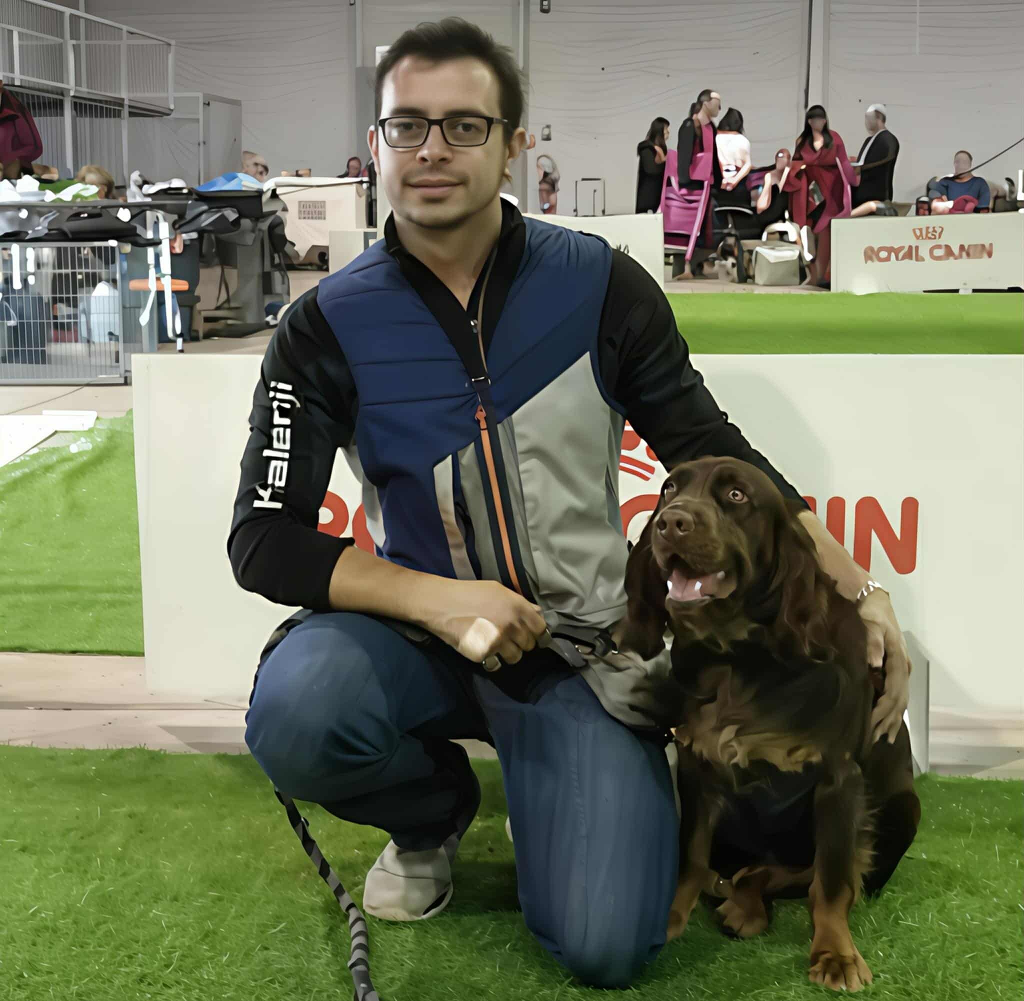 Un homme souriant avec des lunettes, tenant en laisse un chien assis à côté de lui, dans un environnement exposé lors d'un événement canin.